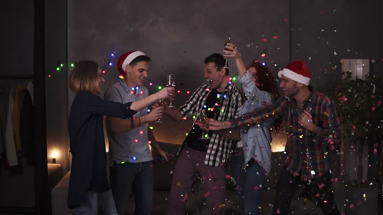 Colorful celebration of Christmas and New year. Festive party, group of five clink glasses with champagne after one guy explodes the confetti stick. Happy celebration together with close friends at home in decorated with garland room