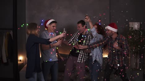 Colorful-celebration-of-Christmas-and-New-year.-Festive-party,-group-of-five-clink-glasses-with-champagne-after-one-guy-explodes-the-confetti-stick.-Happy-celebration-together-with-close-friends-at-home-in-decorated-with-garland-room