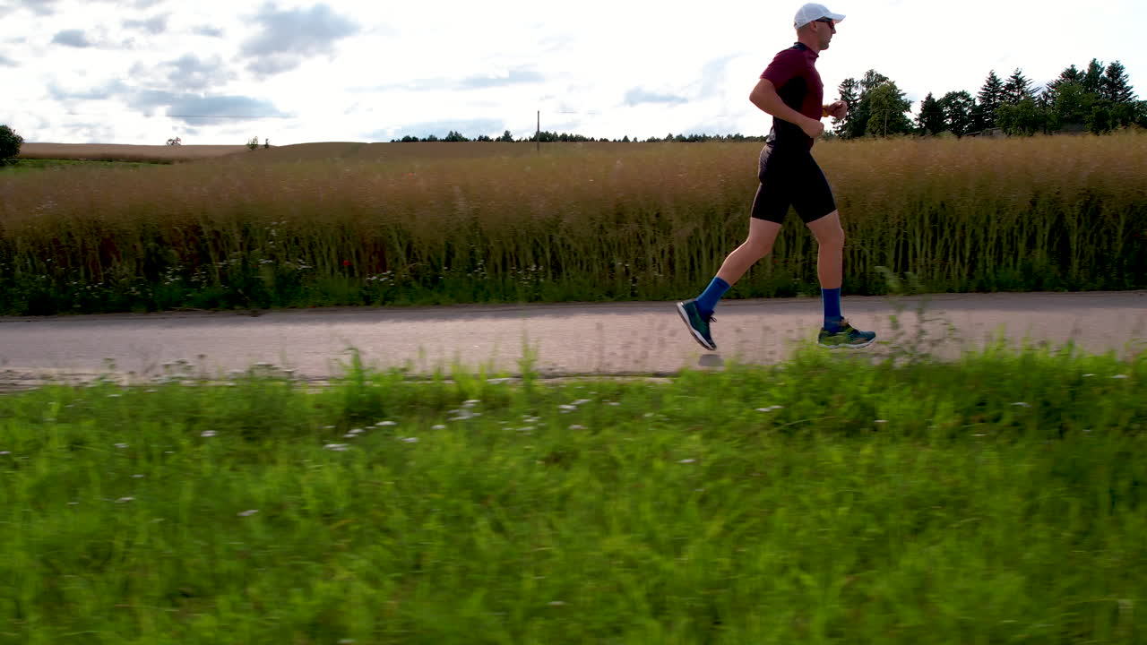 un hombre vestido con ropa deportiva, un sombrero blanco, zapatos de correr y gafas de sol está corriendo por una carretera con un fondo de campos cultivados