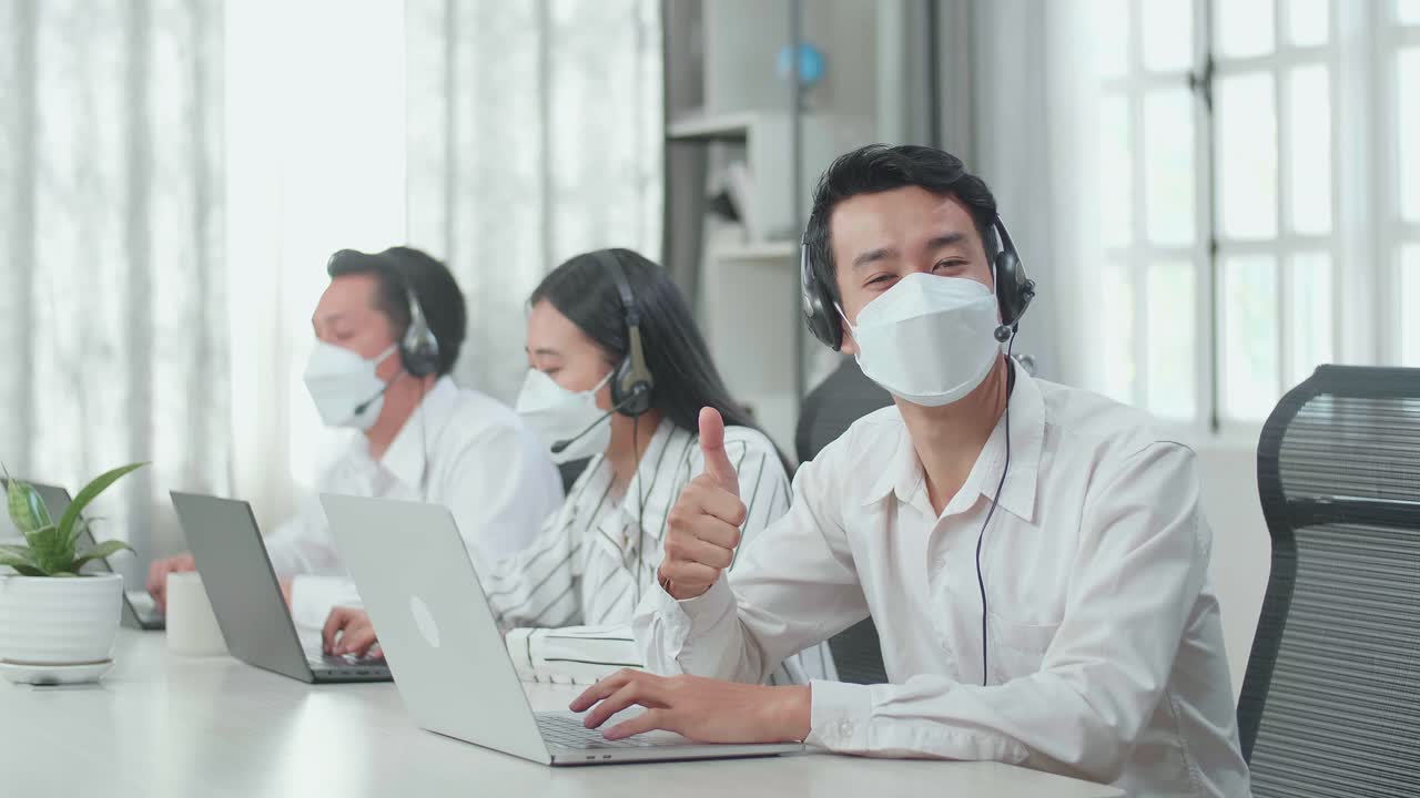 A Man Of Three Asian Call Center Agents Wearing Headset And Mask Looking To Camera From The Computer And Thumbs Up While Two Of His Colleagues Are Speaking With Customer At The Office