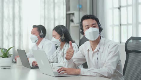 a man of three asian call center agents wearing headset and mask looking to camera from the computer and thumbs up while two of his colleagues are speaking with customer at the office