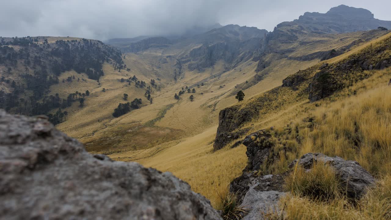 Timelapse statico di una valle di montagna con prati di erba dorata e pendii rocciosi mentre nuvole basse avanzano e la pioggia cade sulle cime distanti