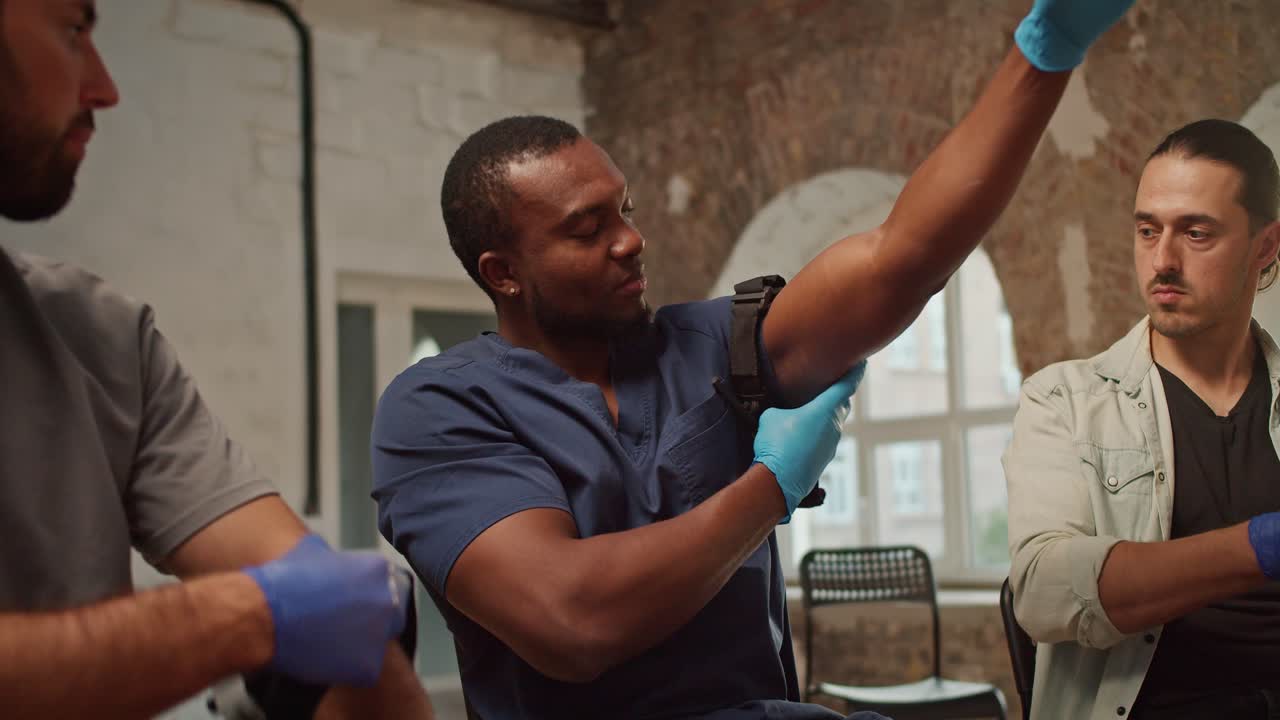 Close-up shooting: A Black male doctor in a blue medical uniform demonstrates a medical harness the technique of tightening the medical straps on the arm during a first aid training