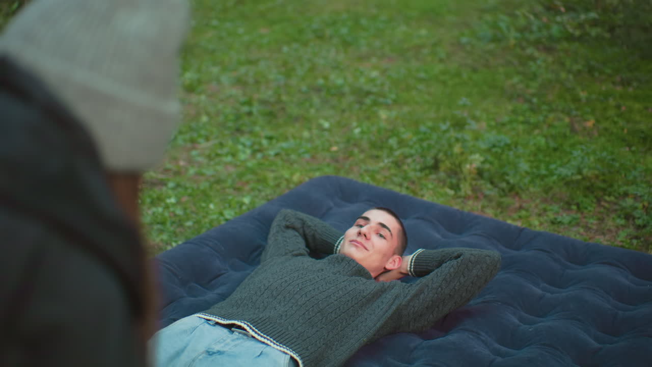 close up rear view of woman standing in foreground while man relaxes on inflated air bed with hands behind head looking at her warmly in grassy forest campsite