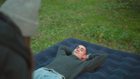 close up rear view of woman standing in foreground while man relaxes on inflated air bed with hands behind head looking at her warmly in grassy forest campsite