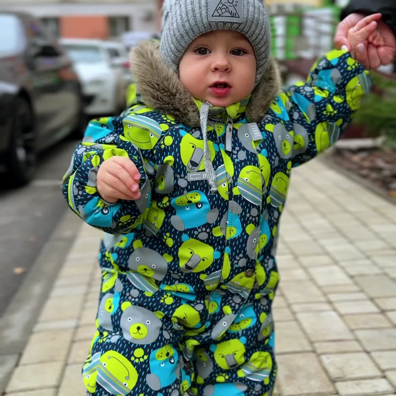 Vertical Screen: Baby wearing warm romper and cap barely starting to walk. Kid is held by the hand wants to go to father