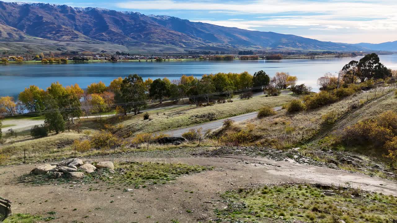Aerial view of Lake Dunstan's serene landscape, featuring a rustic cabin, vibrant autumn foliage, and distant mountains under clear skies