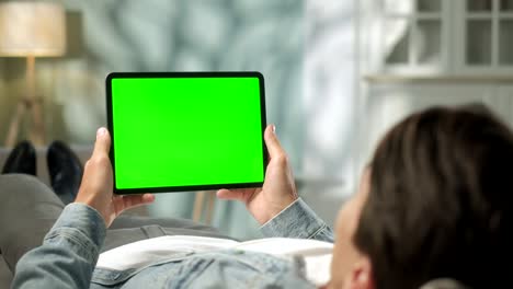 vista desde el hombro del hombre sosteniendo y usando gestos de la mano en la pantalla de maqueta verde computadora tableta digital tendida en un sofá. hombre comprando cosas o navegando tocando en la pantalla central