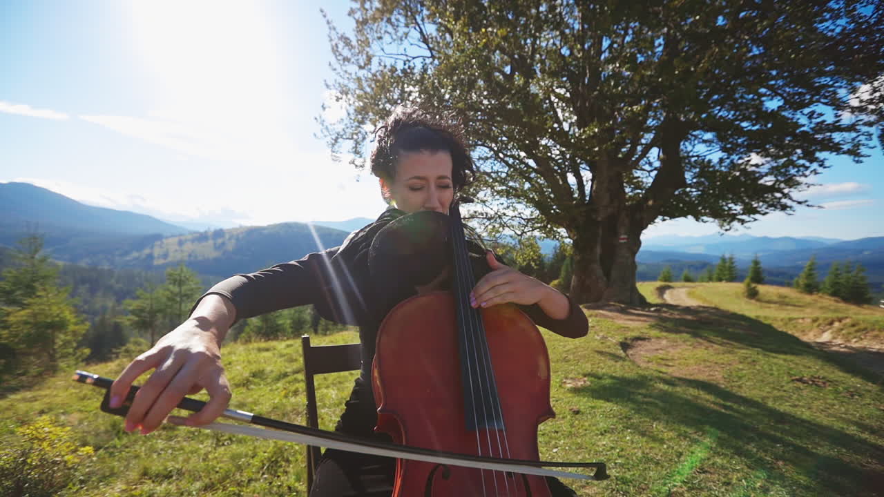 Emotional female musician playing bass-viol in the mountains. Lady in dress sits on the chair playing instrument at the backdrop of beautiful landscape.