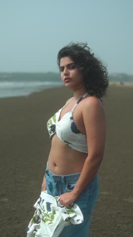 young woman stands on empty beach, wind in hair, holding clothes, calm and thoughtful