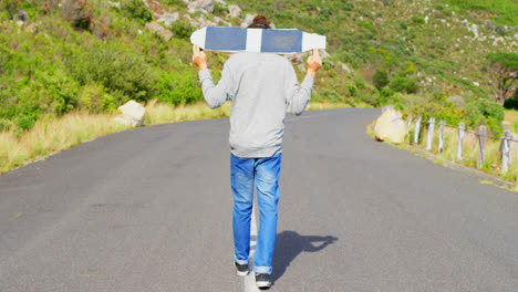 Rear-view-of-cool-young-male-skateboarder-carrying-skateboard-on-shoulder-at-countryside-road-4k