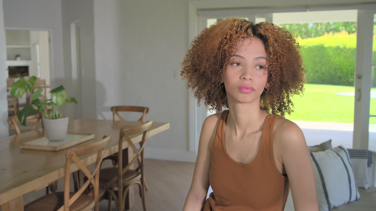 Donna afroamericana che, notando la telecamera, si gira sulla sedia accanto al tavolo vicino alla porta del patio, sorridendo, con spazio per il testo.