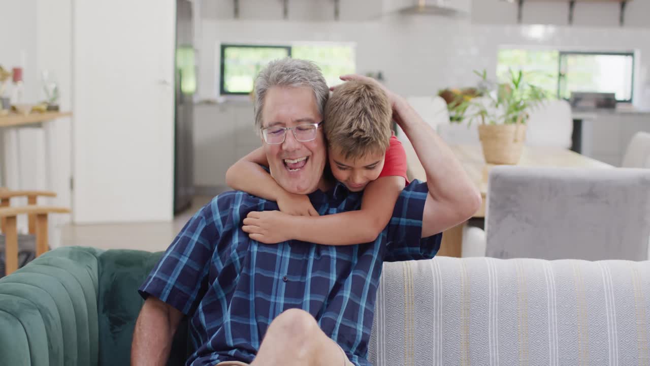 Happy caucasian grandfather and grandson sitting on sofa and embracing, slow motion