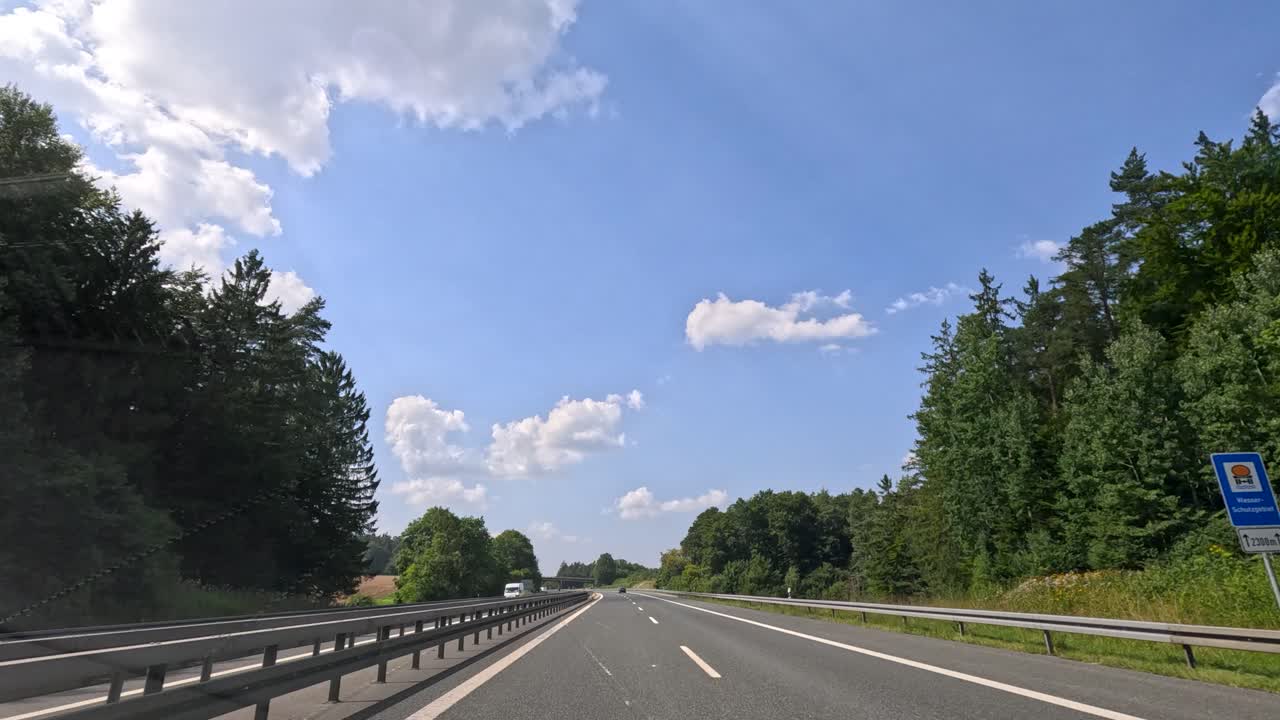 Vehicle travels on sunlit highway near Dunkirk, passing trucks, green forests, blue sky, and clouds