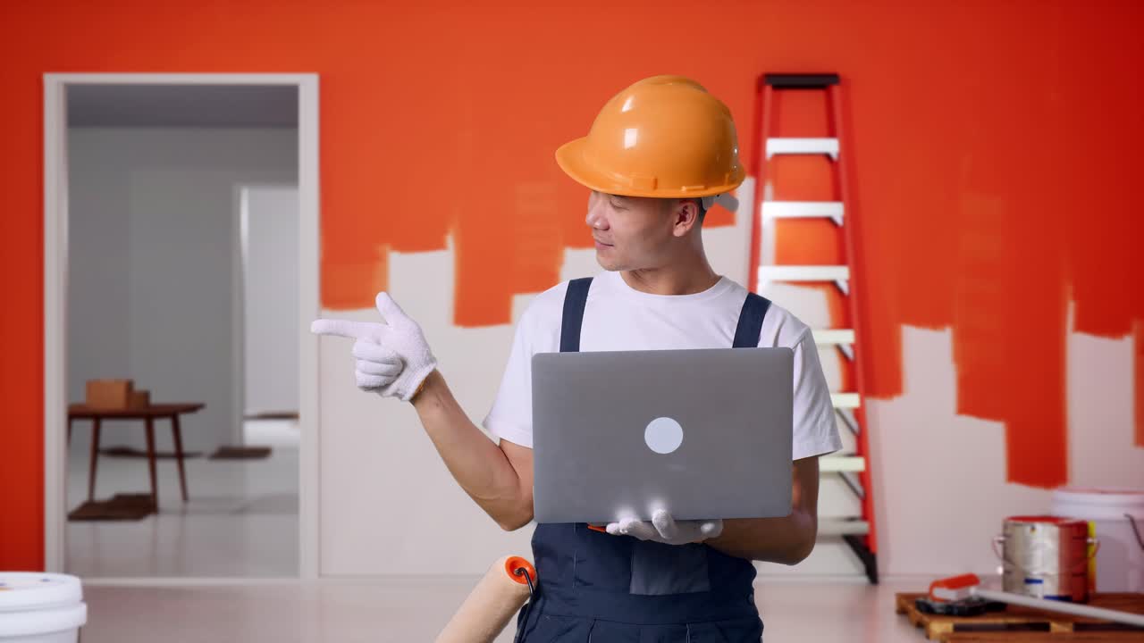 Pittore asiatico che indossa un casco di sicurezza, che usa un laptop e che indica di lato mentre è in piedi con una parete dipinta nella stanza