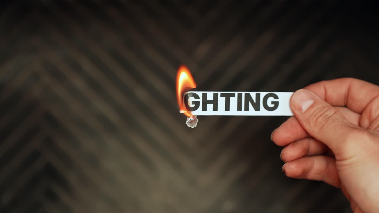 Hand holds a paper strip with the word “LIGHTING” while the edge burns in flame, symbolizing illumination, energy, fire safety, and creative lighting concept. Dark background b-roll