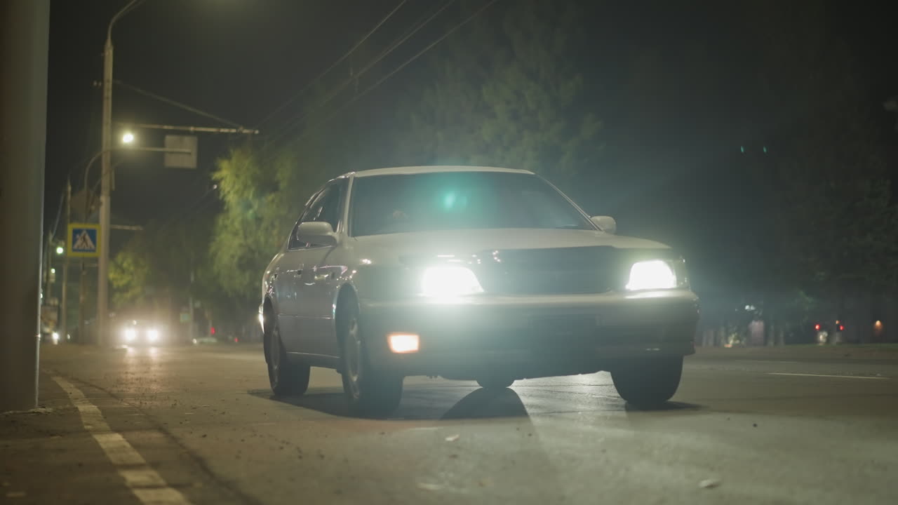 밤거리, 연석으로 다가오는 자동차 전조등, 젖은 아스팔트 위의 가로등 불빛 반사, 멀리 보이는 교통 신호등, 긴장된 도시 분위기, 낮은 앵글에서 느리게 운전하는 모습, 횡단보도 표지판이 보임, 플레어