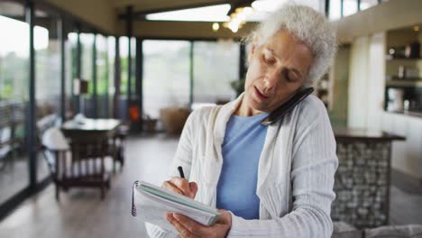 mujer de raza mixta senior hablando en un teléfono inteligente y tomando notas