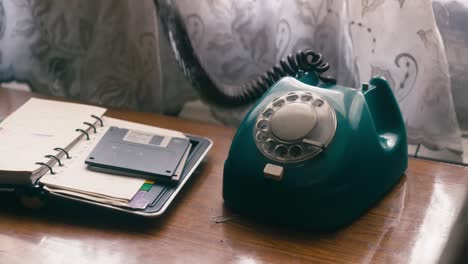 Die-Hand-Einer-Person-Streckt-Sich-Aus,-Um-Ein-Blaugrünes-Telefon-Mit-Wählscheibe-Aufzunehmen,-Das-Neben-Einem-Offenen-Spiralnotizbuch-Auf-Einem-Holztisch-Steht-Und-Eine-Nostalgische-Und-Heitere-Atmosphäre-Schafft