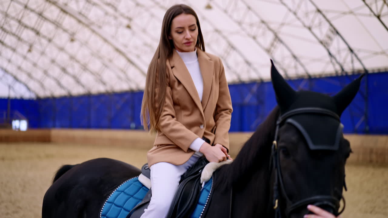 Brunette woman in beige jacket and white pants gets on a horse. Lady is smiling sitting on a black horse. Riding hall at backdrop.