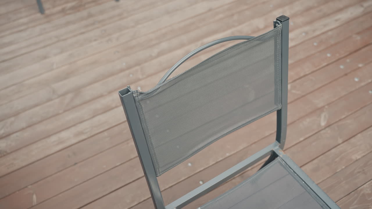 Young caucasian woman sitting on deck, wearing oversized jacket and headphones, adjusting metal mesh chair on wooden floorboards, overhead candid shots capture muted color palette and pensive