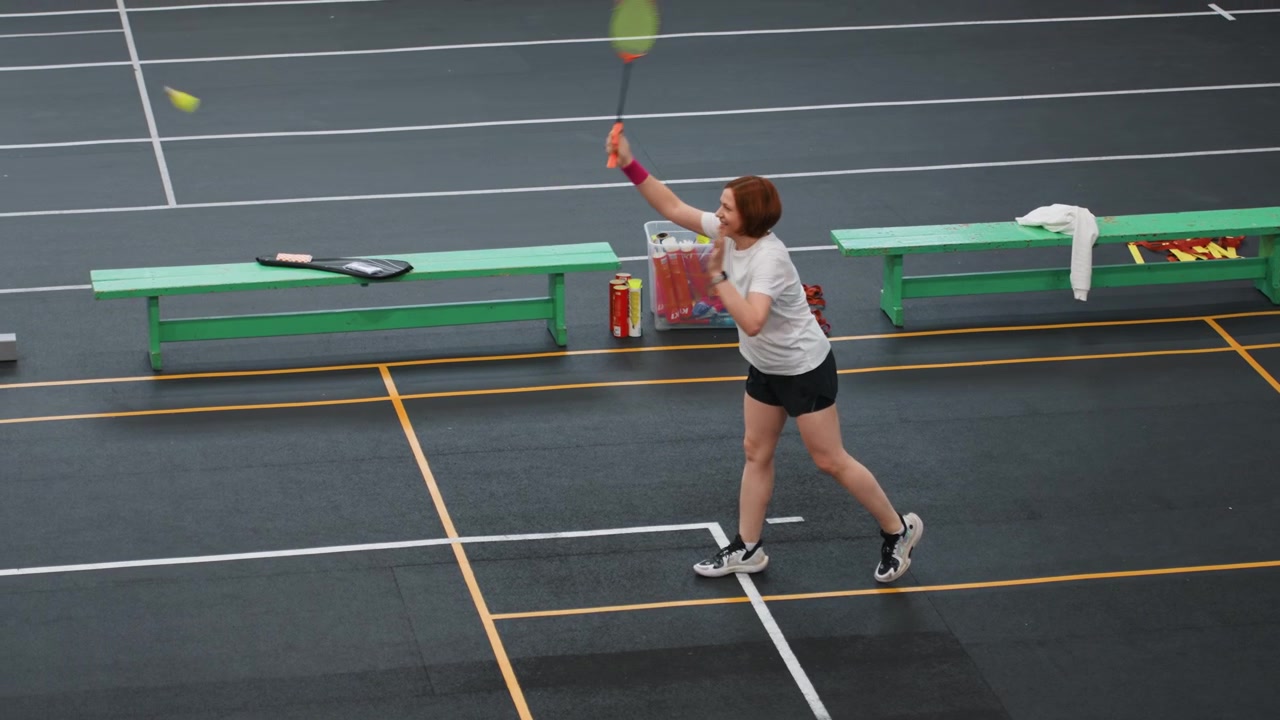 Kvinne spiller badminton i gymsal, klubbstil-rally med forehand- og backhand-slag, hvit t-skjorte og shorts, grønne benker og utstyr ved banekanten, stabilt fotarbeid og leken konkurranseånd