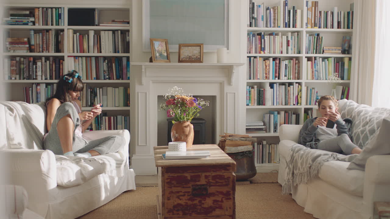 счастливые подростки, использующие смартфоны, расслабляющиеся на диване в гостиной, переписывающиеся в социальных сетях, просматривающие интернет в выходные утром дома