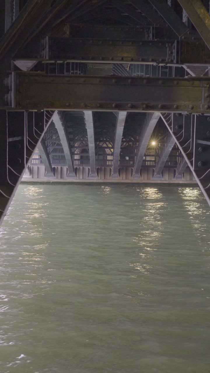 Under a bridge, calm water flows beneath metal beams, serene urban scene