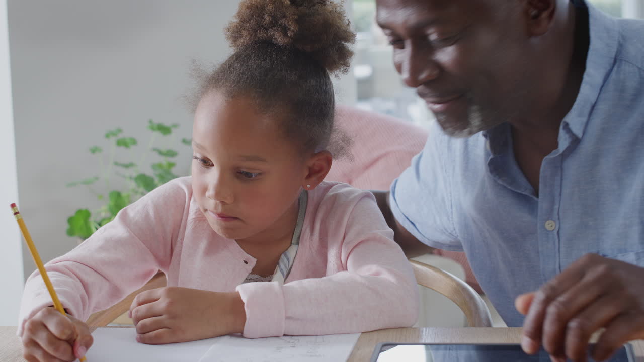Grandfather Helping Granddaughter With Home Schooling Sitting At Table With Digital Tablet