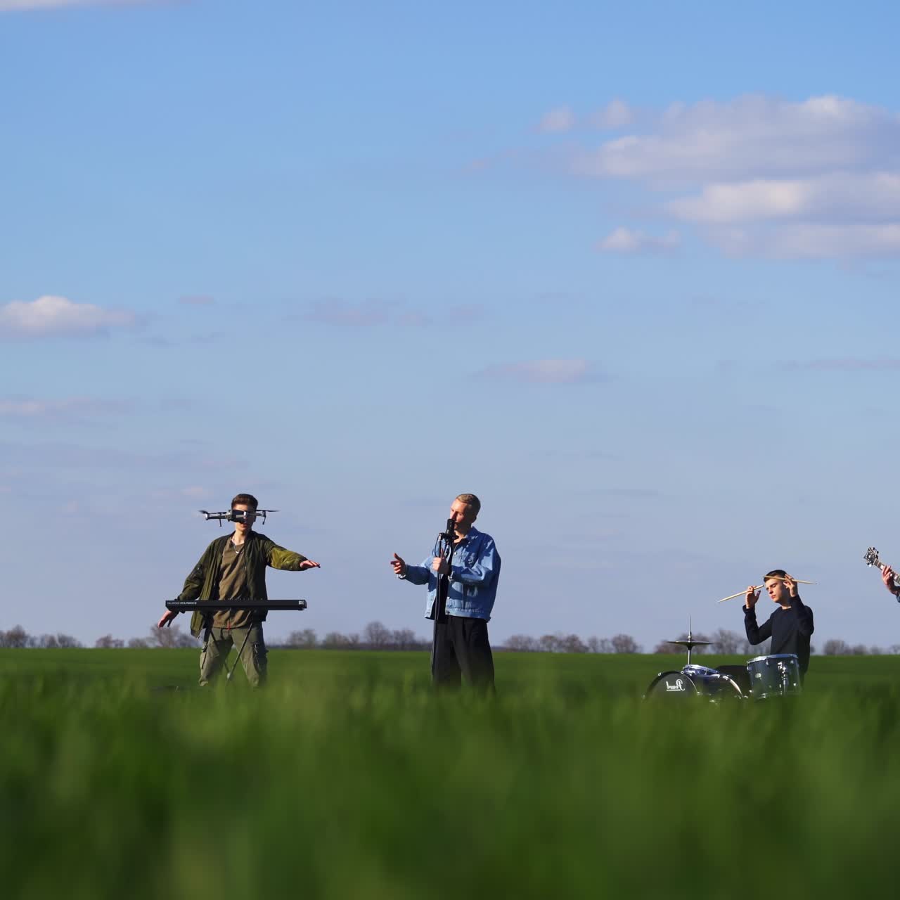 Music band of four guys perform song and instrumental music for drone footage. Gradual view transition from men at backdrop to green grass at foreground