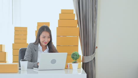 beautiful business asian woman typing on laptop in office with piles of cardboard boxes in background