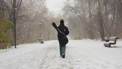 back view of someone carrying a guitar pack while walking on a snowy path in a park, surrounded by bare winter trees and benches dusted with snow