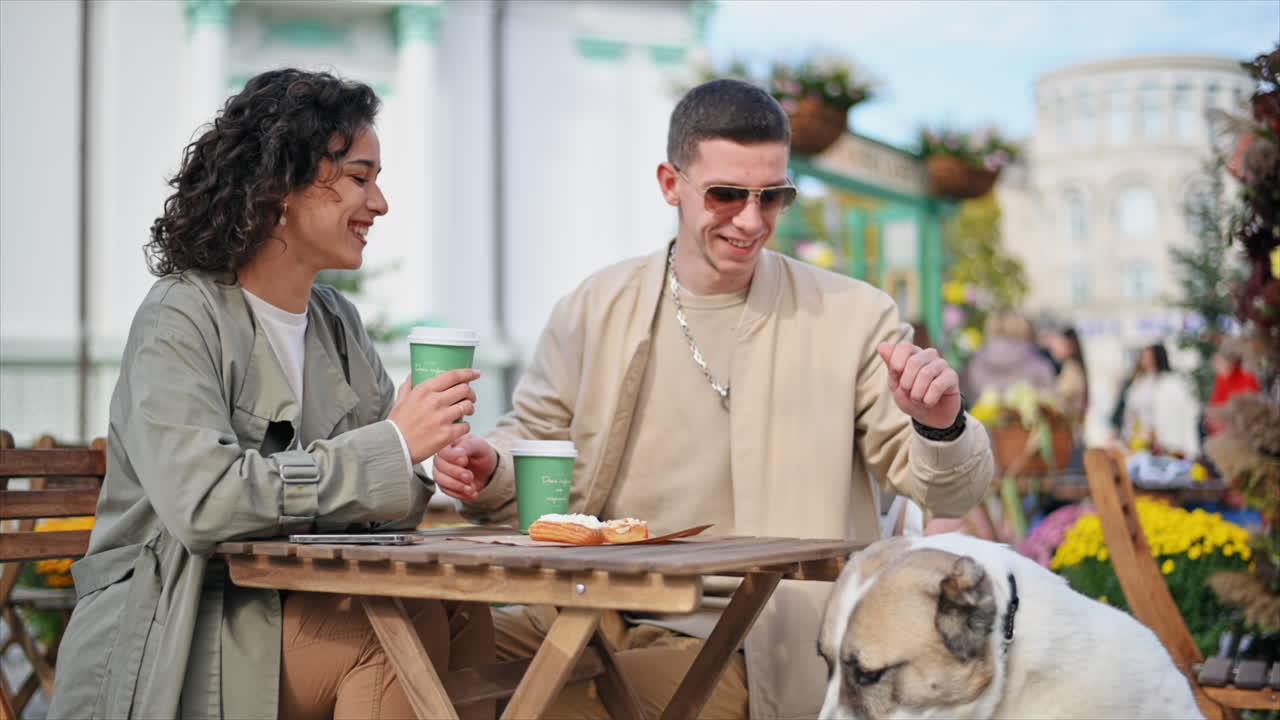 Una coppia felice all'aperto vicino a un bar. Sorridono e danno da mangiare al loro cane, caffè sul tavolo. Atmosfera autunnale. Movimento al rallentatore