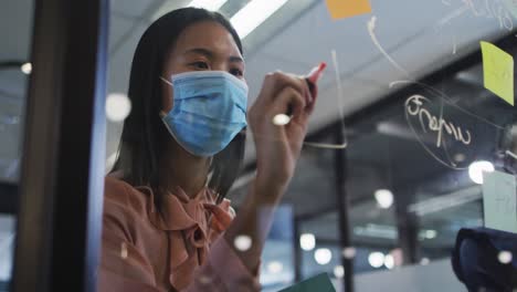 asian woman wearing face mask writing with marker pen on on glass board at modern office