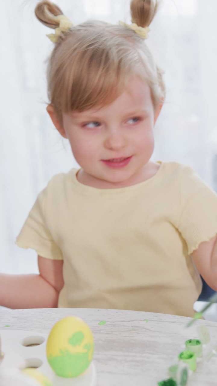 Una bambina con capelli biondi a codini dipinge uova di Pasqua. Si concentra intensamente, tenendo un pennello con cura. La vernice verde copre l'uovo che tiene. La sua espressione mostra una silenziosa concentrazione.