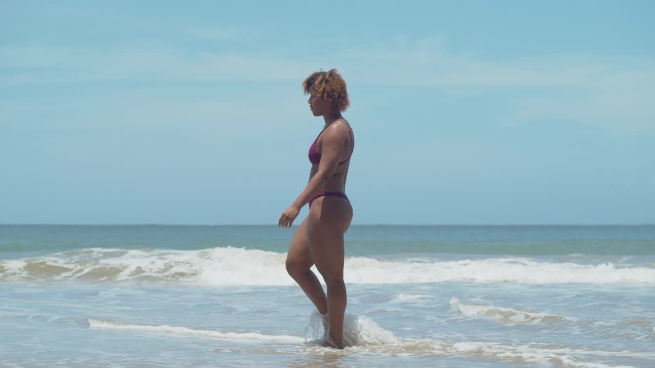 Short curly hair African girl in a bikini wades her feet in the water of the crashing ocean waves