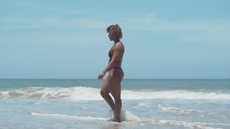 short curly hair african girl in a bikini wades her feet in the water of the crashing ocean waves