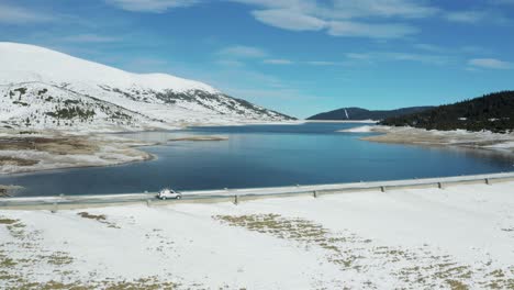 Car-driving-near-a-dam-in-mountain-with-snow-in-winter,-aerial-side-shot