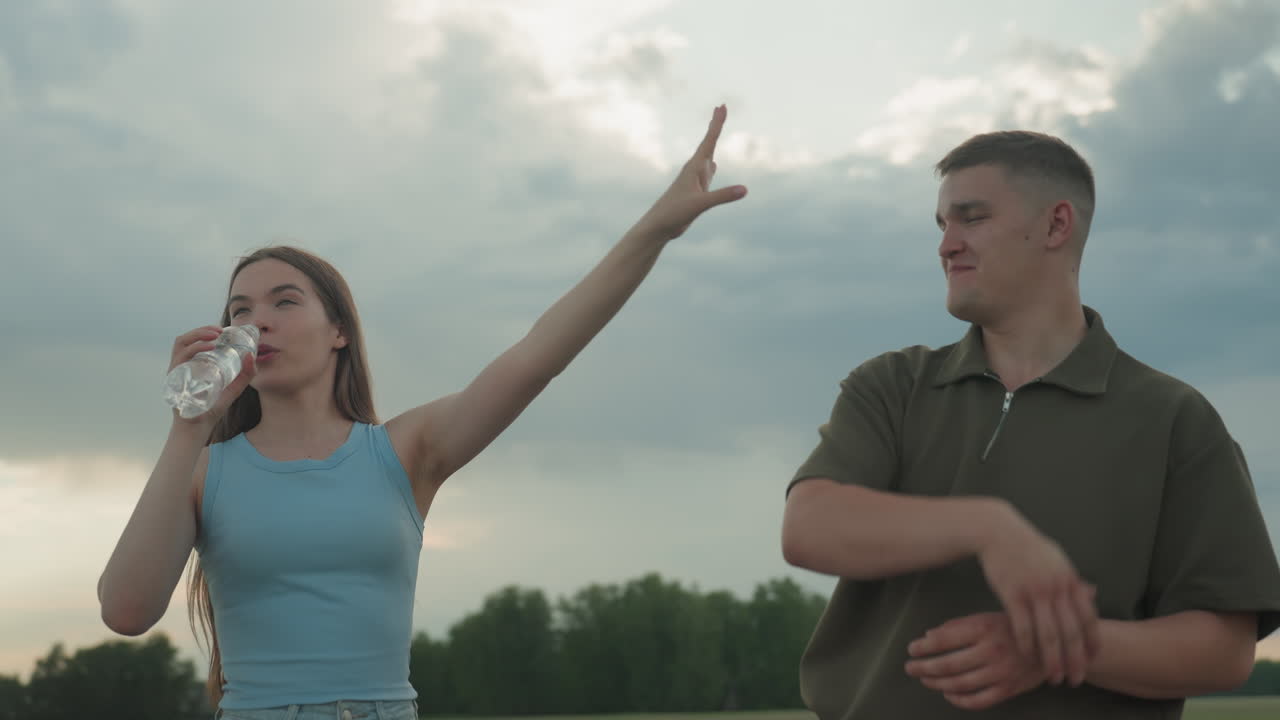 brother reaches for water while sister drinks from plastic bottle then playfully pushes his hand away on deserted country road at sunset sky capturing sibling playful moment next to parked car