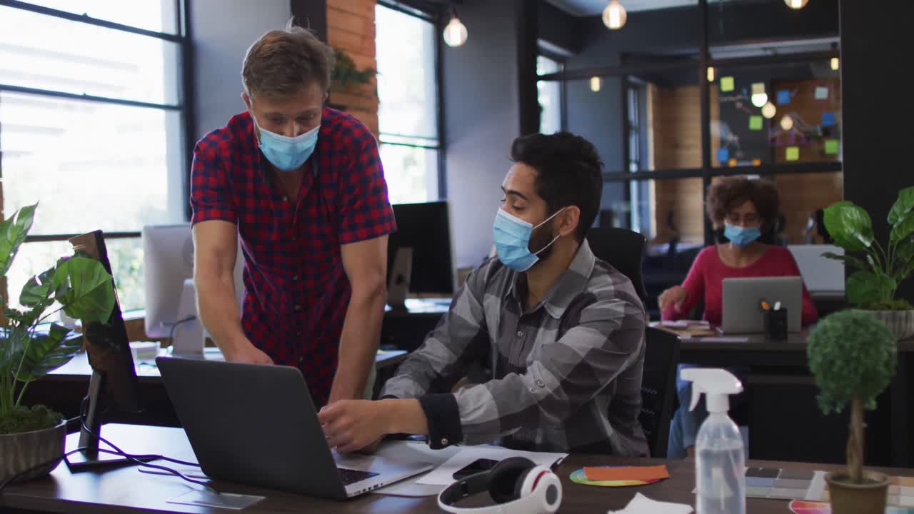 Diverse male colleagues wearing face masks discussing over laptop at modern office