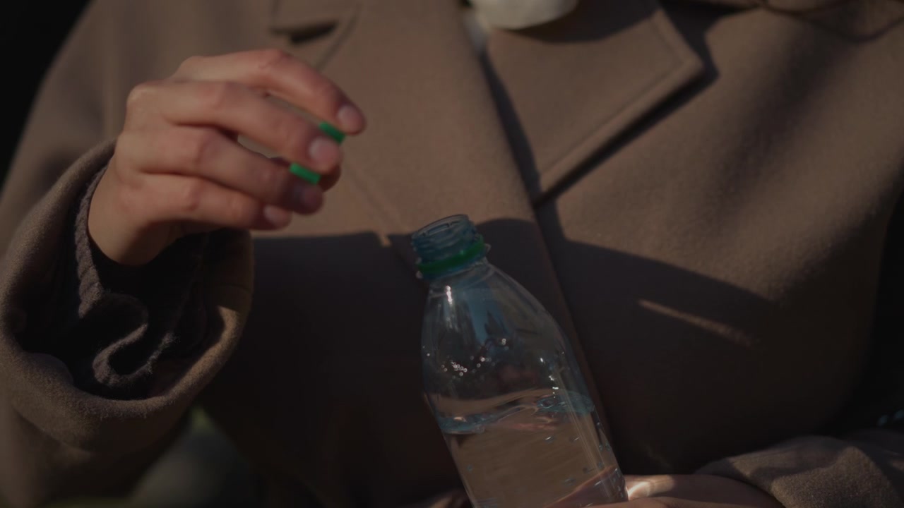 Close-up seorang wanita berjaket cokelat sedang membuka botol air plastik bertutup hijau, tangan terlihat jelas. Cahaya alami yang lembut menyoroti detail tekstur pada kain dan cairan bening.