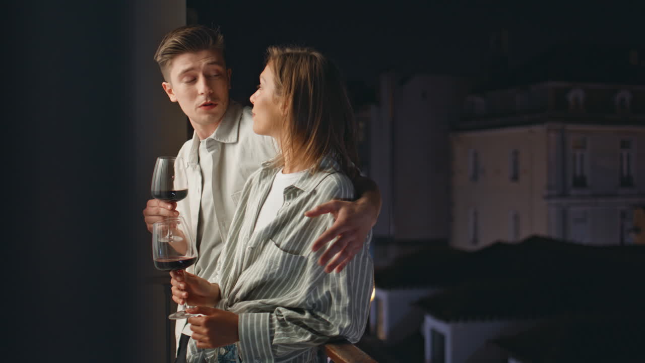 Relaxed couple talking balcony at night. Smiling man embracing girl holding wine