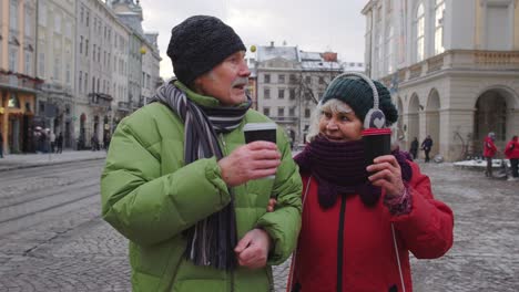 senior old tourists grandmother grandfather walking, drinking hot drink mulled wine in city center