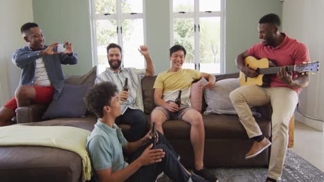 happy diverse male friends playing guitar and talking in living room