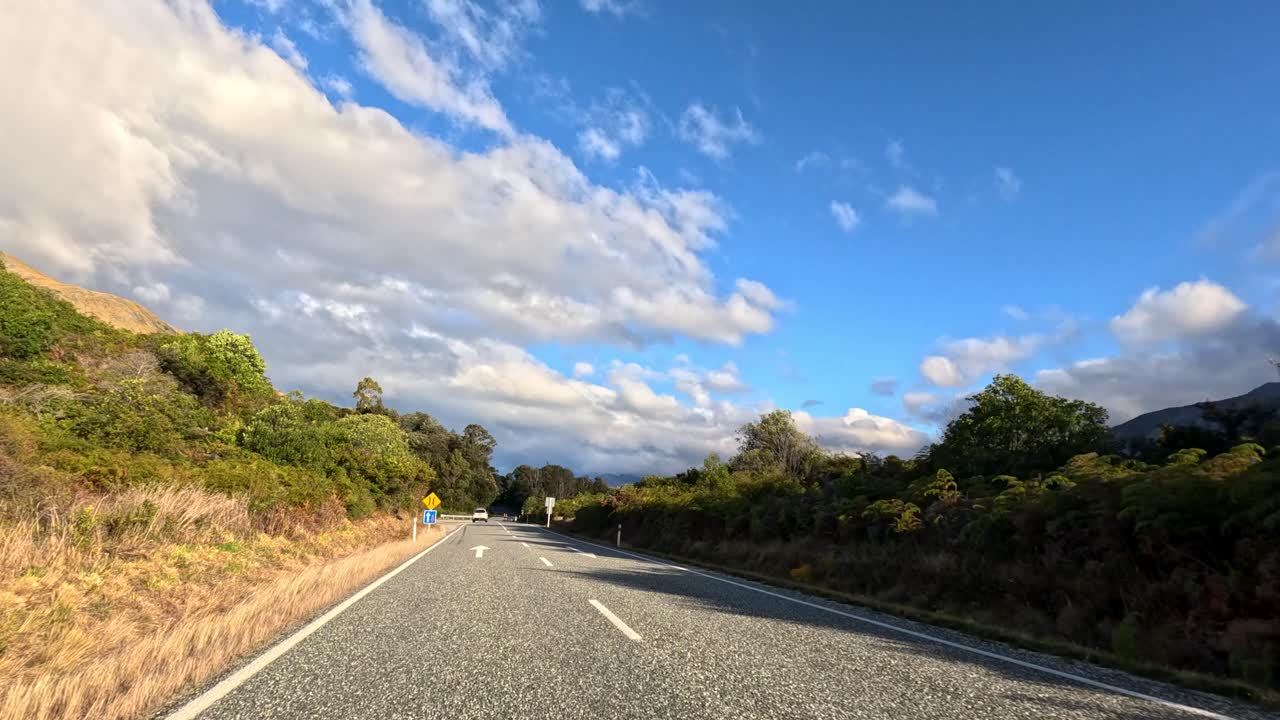 Vehicle travels winding mountain road, passing dry grass, green bushes, under bright afternoon sunlight
