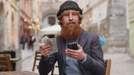 happy young man counting money dollar cash use smartphone calculator app in urban city street