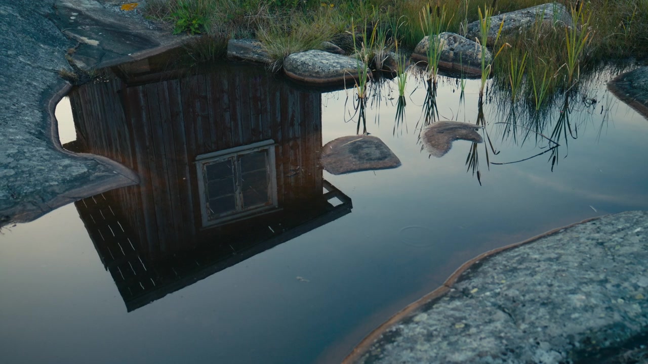 Riflesso astratto di una rimessa per barche in legno stagionato specchiato in una tranquilla pozza di marea, circondata da liscio granito costiero e vegetazione litoranea. Paesaggio acquatico scandinavo pacifico con texture naturali