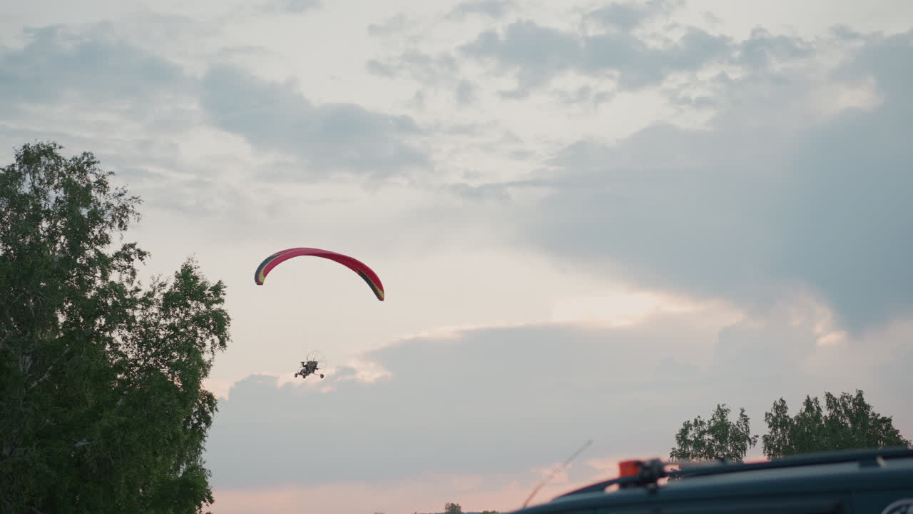 powered paraglider with red canopy soars above treetops during golden hour against dramatic sky with scattered clouds and soft light