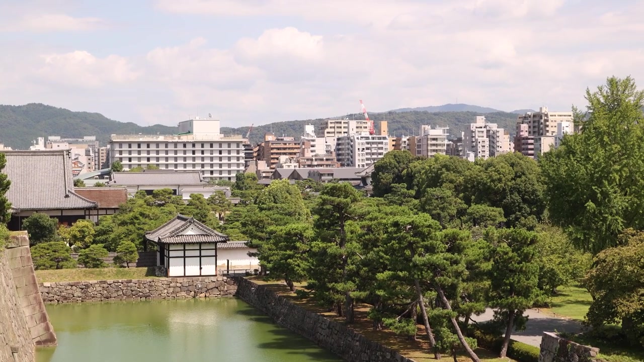 Vista dall'alto delle storiche mura in pietra, del fossato e dei rigogliosi giardini del Castello di Nijo a Kyoto, in Giappone, sullo sfondo del moderno paesaggio urbano