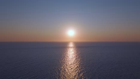 drone drifts backward from deep over the ocean toward the beach, capturing the glowing sunset and its stunning reflection on the water’s surface in a peaceful, mesmerizing golden hour moment.